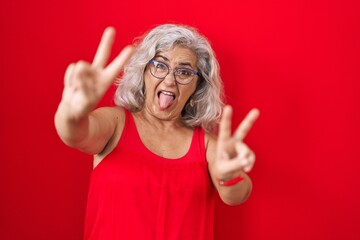 Middle age woman with grey hair standing over red background smiling with tongue out showing...