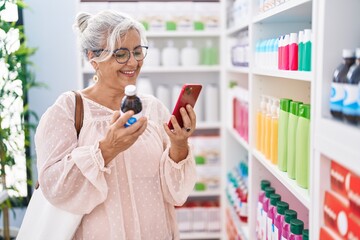 Middle age grey-haired woman customer using smartphone holding medicine bottle at pharmacy