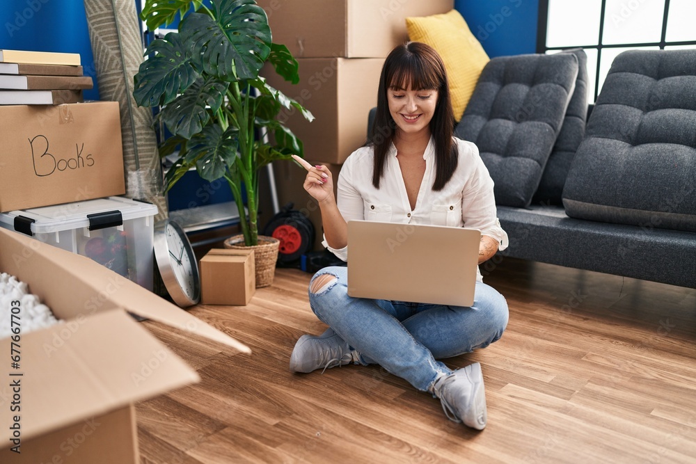 Sticker young brunette woman sitting on the floor with laptop at new home smiling happy pointing with hand a