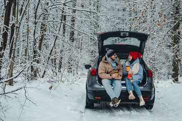 In a snowy winter forest, a couple drinks coffee in their trunk