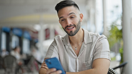 Obraz premium Young hispanic man using smartphone sitting on table smiling at coffee shop terrace