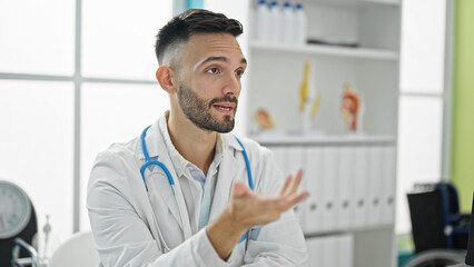 Young hispanic man doctor sitting on table speaking at the clinic