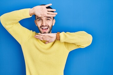 Hispanic man standing over blue background smiling cheerful playing peek a boo with hands showing...