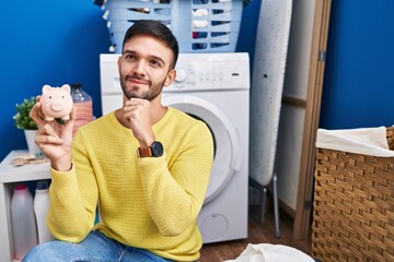 Hispanic man doing laundry holding piggy bank serious face thinking about question with hand on...