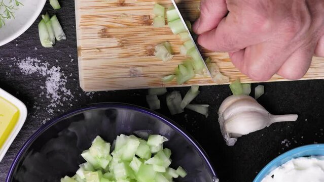 The chef is dicing cucumbers into small cubes to prepare Bulgarian Snezhanka salad, consisting of strained yogurt, olive oil, garlic, and dill herb.