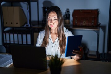 Young brunette woman working at the office at night puffing cheeks with funny face. mouth inflated with air, crazy expression. - obrazy, fototapety, plakaty