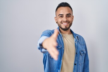 Young hispanic man standing over isolated background smiling friendly offering handshake as greeting and welcoming. successful business.