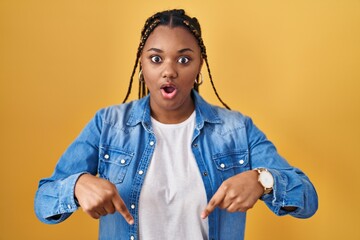 African american woman with braids standing over yellow background pointing down with fingers...
