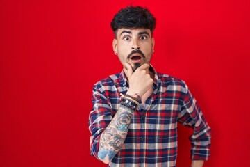 Young hispanic man with beard standing over red background looking fascinated with disbelief, surprise and amazed expression with hands on chin