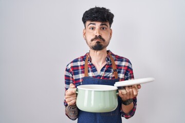 Young hispanic man with beard wearing apron holding cooking pot puffing cheeks with funny face. mouth inflated with air, catching air. - obrazy, fototapety, plakaty