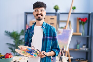 Young hispanic man artist holding paintbrush and palette at art studio