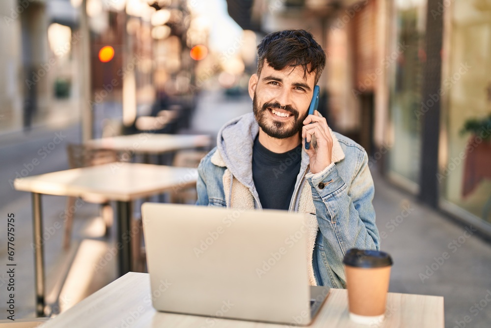 Poster young hispanic man using laptop talking on smartphone sitting on table at coffee shop terrace