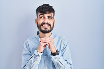 Young hispanic man with beard standing over blue background laughing nervous and excited with hands on chin looking to the side