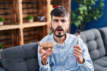 Young hispanic man with beard eating healthy whole grain cereals in shock face, looking skeptical and sarcastic, surprised with open mouth