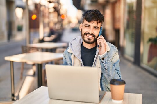 Young hispanic man using laptop talking on smartphone sitting on table at coffee shop terrace