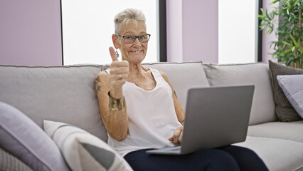 Cheerful, grey-haired senior woman sitting on sofa at home, letting good vibes flow while using...