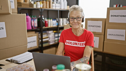 Senior grey-haired woman volunteer using laptop and headphones smiling at charity center