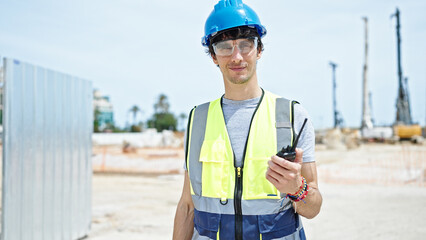 Young hispanic man architect holding walkie-talkie smiling at construction place