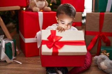 Fototapeta na wymiar Adorable hispanic toddler unpacking christmas gift sitting on floor at home
