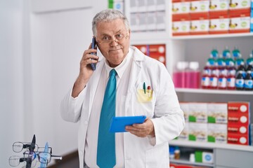Middle age grey-haired man pharmacist using touchpad talking on smartphone at pharmacy