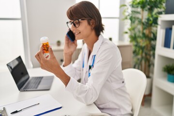 Young beautiful hispanic woman doctor talking on smartphone holding pills bottle at clinic