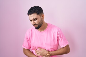 Hispanic young man standing over pink background with hand on stomach because indigestion, painful illness feeling unwell. ache concept.