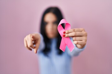 Hispanic woman holding pink cancer ribbon pointing with finger to the camera and to you, confident gesture looking serious - obrazy, fototapety, plakaty