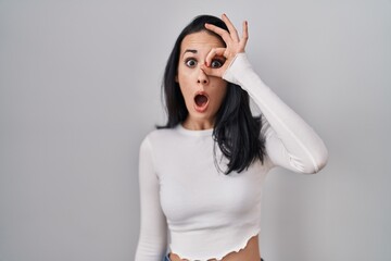 Hispanic woman standing over isolated background doing ok gesture shocked with surprised face, eye looking through fingers. unbelieving expression.