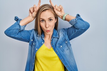 Young blonde woman standing over blue background doing funny gesture with finger over head as bull horns - obrazy, fototapety, plakaty
