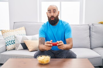 Young hispanic man with beard and tattoos playing video game sitting on the sofa afraid and shocked with surprise and amazed expression, fear and excited face.