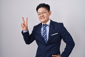 Young asian man wearing business suit and tie smiling looking to the camera showing fingers doing victory sign. number two.