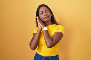 Young indian woman standing over yellow background sleeping tired dreaming and posing with hands together while smiling with closed eyes.