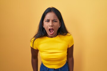 Young indian woman standing over yellow background angry and mad screaming frustrated and furious,...
