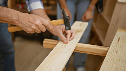 Two skilled carpenters, a dynamic duo, driving a nail into a wood plank at a lively carpentry workshop. joiner and handyman together hammering, shaping the timber into bespoke furniture
