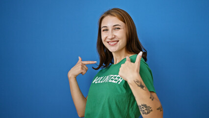 Young woman pointing herself to volunteer uniform smiling over isolated blue background