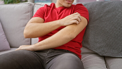 Portrait of a young, handsome hispanic man sitting on sofa at home, scratching itchy arms - a...