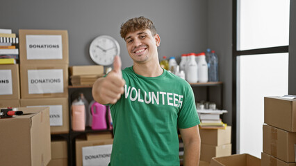 Smiling young hispanic man confidently volunteering at charity center, giving thumbs up gesture in...
