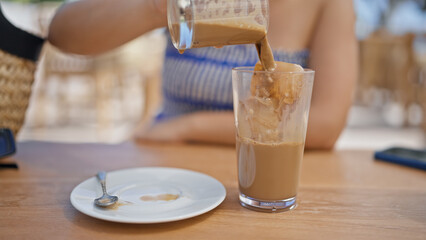 Young hispanic woman pouring coffee into glass with ice at sunny restaurant terrace