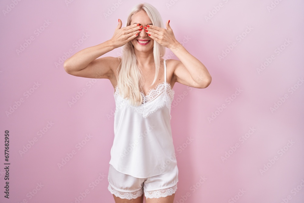 Sticker Caucasian woman wearing pajama wearing pink background covering eyes with hands smiling cheerful and funny. blind concept.