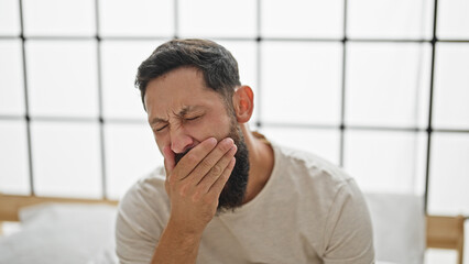 Young hispanic man sitting on bed yawning at bedroom