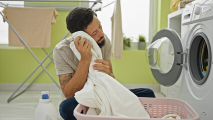 Young hispanic man washing clothes touching clean towel with face at laundry room