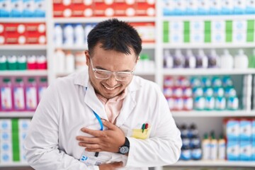 Chinese young man working at pharmacy drugstore smiling and laughing hard out loud because funny crazy joke with hands on body.