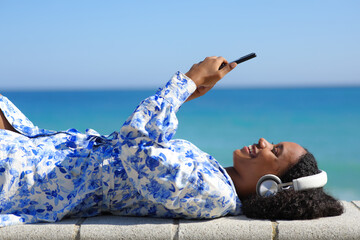 Happy black woman lying on the beach listening audio on phone - obrazy, fototapety, plakaty