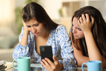 Two sad women checking bad news on cell phone