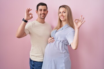 Fototapeta premium Young couple expecting a baby standing over pink background smiling positive doing ok sign with hand and fingers. successful expression.