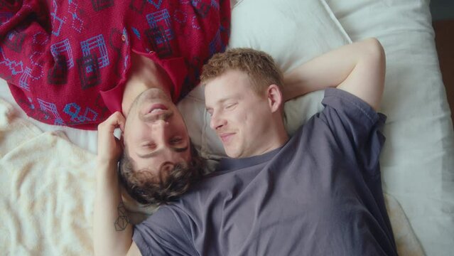 Young happy gay couple chatting, smiling and kissing while lying on bed at home and enjoying romantic moments together. Directly above view