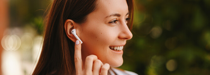 Banner photo of young joyful woman using earpods outdoor, wireless headphones.