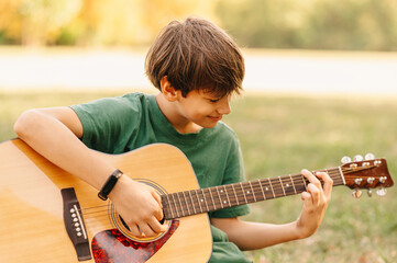 Young boy sitting on grass in park and practicing at acoustic guitar.