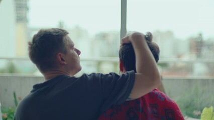 Affectionate gay man stroking hair of beloved boyfriend, then kissing him with love while sitting together by the window at home. View from behind
