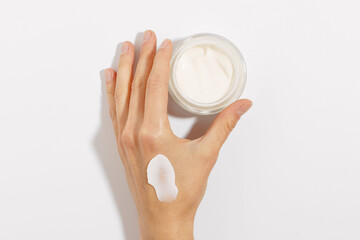 female hand with a smear of white cream holding a glass jar of moisturizer on a white isolated background. concept of beauty products. mockup for your design